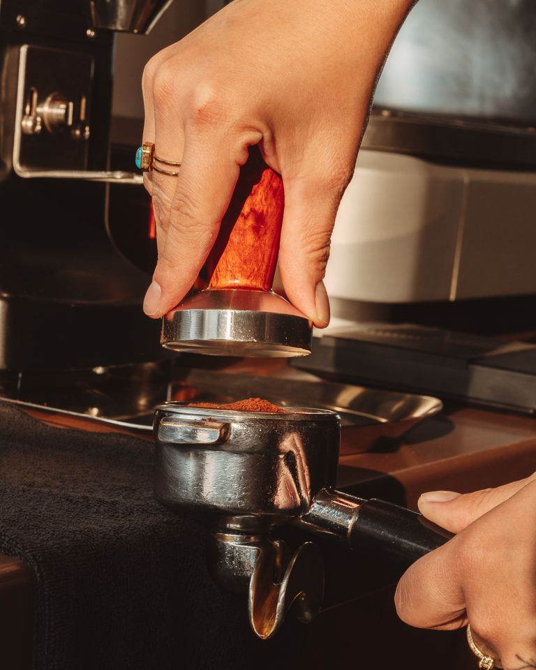 A hand expertly tamping coffee grounds into a portafilter, set in a warm light setting, hinting at the artisan coffee-making process.