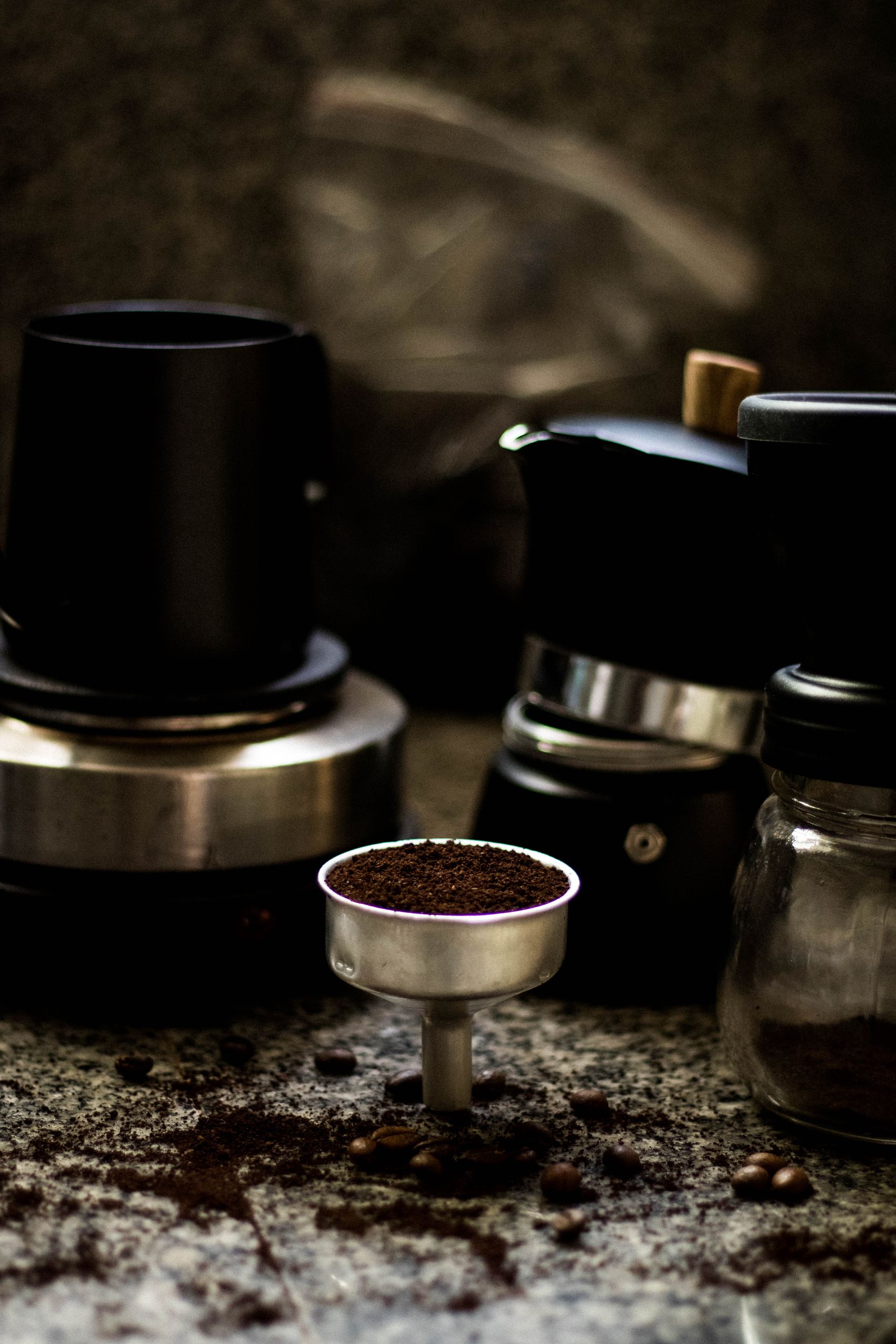 Dark, moody still life of espresso equipment with coffee grounds on textured surface.