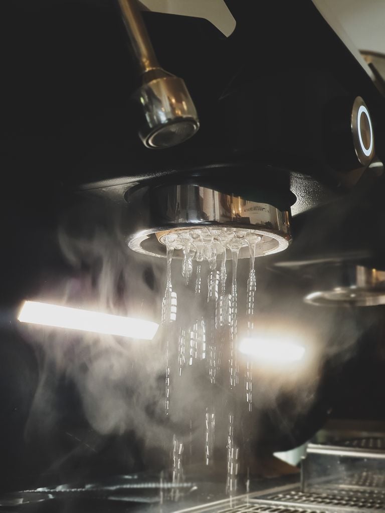 Steam and hot water pour from an espresso machine group head, creating a moody atmospheric effect.